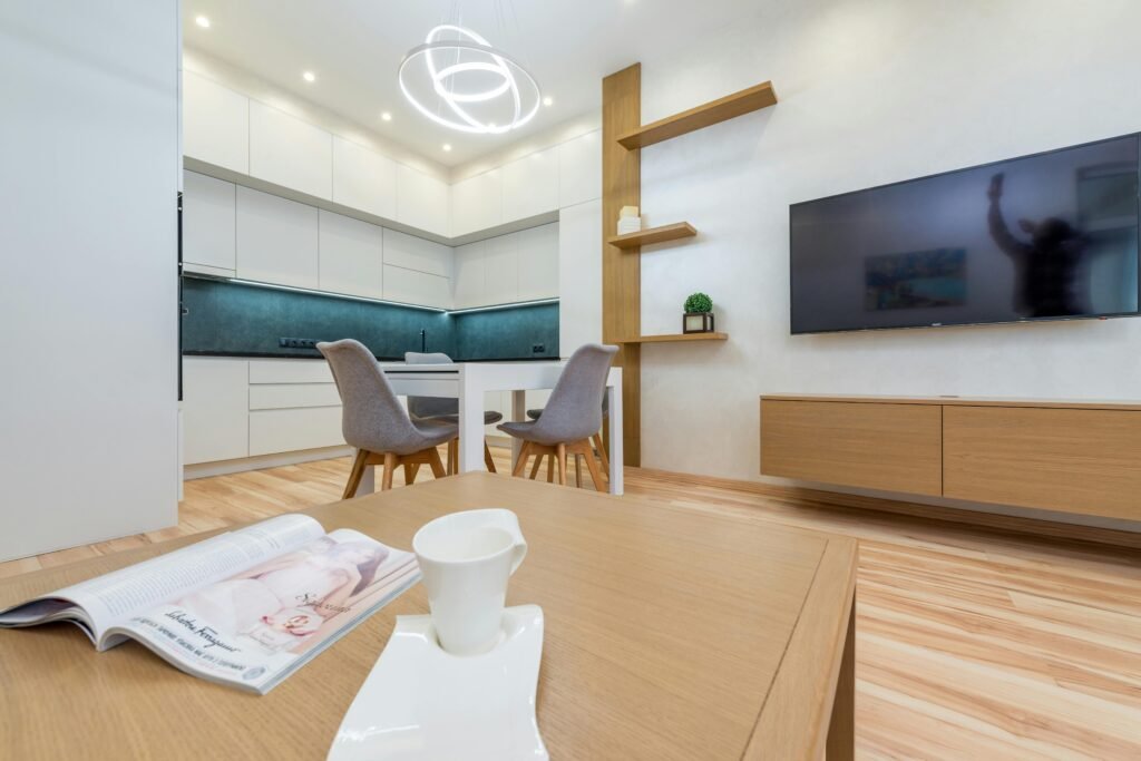 Interior of modern light room with table with mug and magazine in front of TV on wall next to chairs and open kitchen with cabinets and shelves